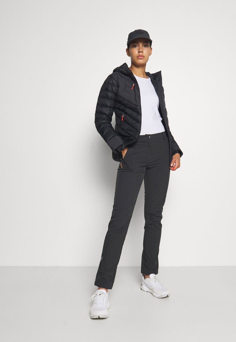 Zwarte pufferjack met rode accenten, witte t-shirt en zwarte broek. Schoenen zijn witte sportieve schoenen. Model staat zelfverzekerd tegen een neutrale achtergrond.