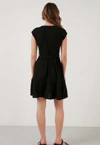 Svart klänning med utsvängd kjol och korta ärmkragar, texturerad tyg, åtstramad midja och volang nederkant. Sedd bakifrån.