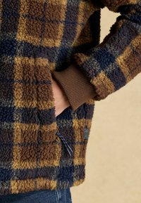 Fleece jacket featuring a brown and navy checkered pattern, ribbed cuffs, and a zippered pocket. Soft texture and relaxed fit.