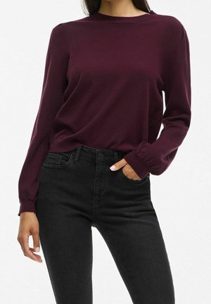 Kobieta w długim rękawie ciemnofioletowego swetra i czarnych, wysokich dżinsach z jedną ręką w kieszeni na białym tle.