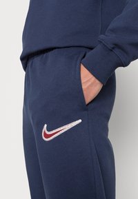 Mörkblå sweatpants med en mjuk textur, som har en broderad vit och vinröd Nike-logotyp på vänster lårområde. Elastisk midja med fickor.