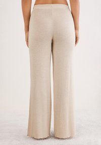 Pantalons évasés beige clair en tissu doux et texturé avec une taille lisse. L'ourlet touche le sol, mettant en valeur la coupe décontractée.