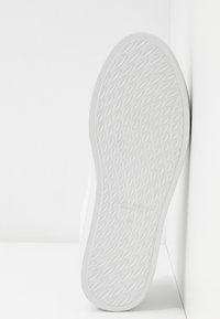 Semelle de basket blanche avec un grip texturé en forme de losanges. Design minimaliste avec un bord lisse et un logo embossé près du talon.