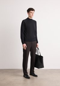 Schwarzer Langarmpullover mit hohem Kragen, kombiniert mit dunkelbraunen, maßgeschneiderten Hosen und schwarzen Lederschuhen. Hält eine schwarze Tragetasche.