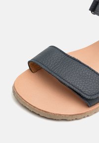 Mörkblå lädersandal med en texturerad rem, ljusbeige fotbädd och en flexibel gummisula. Närbild av design detaljer.