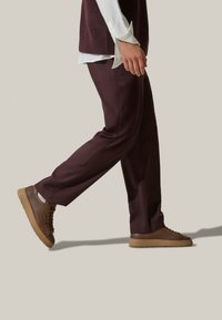 Pantaloni taglio sartoriale bordeaux con una texture liscia abbinati a sneakers marroni con suola piatta e lacci minimal, mostrati in profilo laterale.