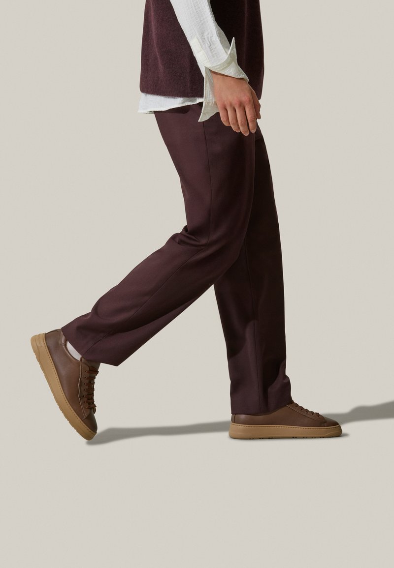 Pantaloni taglio sartoriale bordeaux con una texture liscia abbinati a sneakers marroni con suola piatta e lacci minimal, mostrati in profilo laterale.