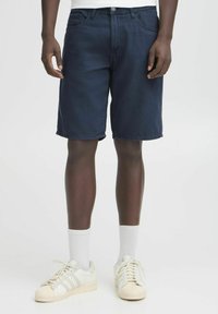 Dark blue denim shorts with a relaxed fit, five pockets, and a zip fly. Worn with white socks and white sneakers featuring three stripes.