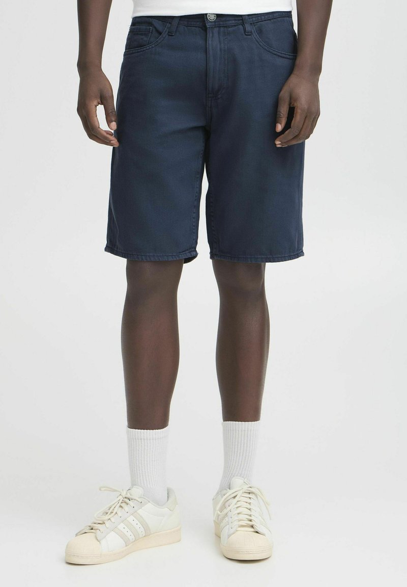 Dark blue denim shorts with a relaxed fit, five pockets, and a zip fly. Worn with white socks and white sneakers featuring three stripes.