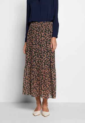 Frau trägt einen dunkelgrünen Maxi-Rock mit kleinem Pfirsichblumenmuster, eine marineblaue Langarmbluse und cremefarbene spitze Schuhe.