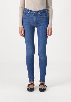 Osoba ubrana w obcisłe niebieskie jeansy, beżowy prążkowany longsleeve i czarne płaskie buty z klamrami, stojąca na jasnym, jednolitym tle.