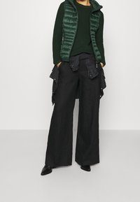 Gilet imbottito verde sopra un maglione testarossa verde scuro, abbinato a pantaloni larghi neri e scarpe nere a punta.
