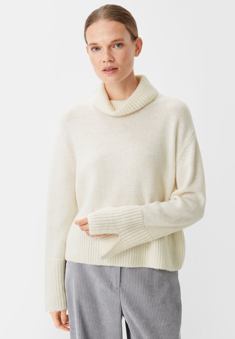 Beige gestrickter Pullover mit einem Rollkragen, weiten Ärmeln und gerippte Bündchen. Weiche Textur mit einer lockeren Passform, kombiniert mit grau gestreiften Hosen.