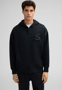 Sweat à capuche zippé noir en tissu doux, avec une poche frontale et un logo "S" texturé sur la poitrine. Il a une coupe décontractée et une capuche.