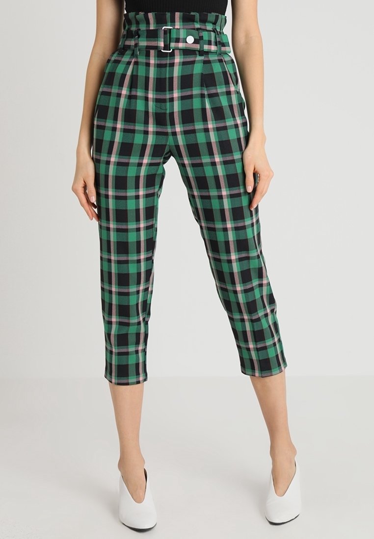 Femme portant un pantalon court taille haute à motif écossais vert, noir et rose, une ceinture assortie et des chaussures blanches à talons.