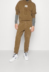 Sudadera de algodón marrón con cordones, pantalones ajustados a juego y zapatillas blancas. Presenta parches con logos y una textura desgastada.