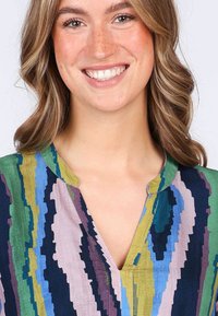 Bunt gemusterte Bluse mit vertikalen Zickzackmustern in verschiedenen Grüntönen, Blau, Lila und Pink, versehen mit einem kleinen V-Ausschnitt.