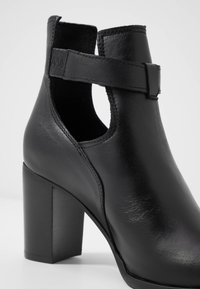 Bottine en cuir noir à talon épais. Comprend des côtés découpés et une bride ajustable. Texture lisse avec des détails de couture subtils.