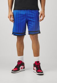 Kék kosárlabda nadrág fekete függőleges csíkokkal, oldalsó hasítékokkal, fekete és piros magas szárú sportcipővel párosítva. Fehér zoknik láthatók.