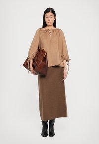 Braune oversized Bluse mit Bindedetails, kombiniert mit einem braunen Strickrock. Das Model hält eine dunkelbraune Tasche. Schwarze kniehohe Stiefel vervollständigen den Look.