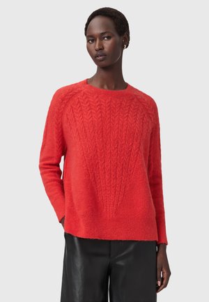Femme portant un pull en maille torsadée rouge vif et un pantalon en cuir noir, se tenant avec une main dans la poche contre un fond uni.