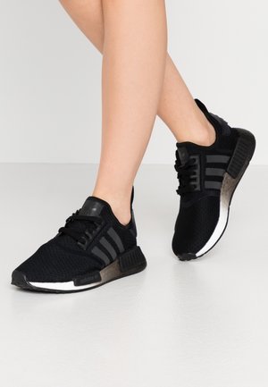 Nogi w czarnych butach sportowych Adidas z dzianą teksturą, trzema ciemnymi paskami i białymi podeszwami, stojące na jasnoszarej podłodze na jednolitym tle.