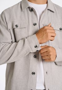 Ljust grå herringbone skjortjacka med svarta knappar, två bröstfickor, långa ärmar och rak krage, lager på lager över en vit skjorta.