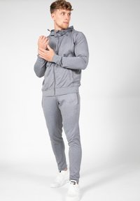 Grijze sportieve trainingsoutfit gemaakt van glad, rekbaar materiaal. Bevat een jack met rits en capuchon en aansluitende broek, gecombineerd met witte sneakers.