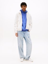 Witte fleecejas over een blauwe hoodie, lichtblauwe distressed jeans en witte sneakers met blauwe accenten. Casual outfit met zachte texturen.