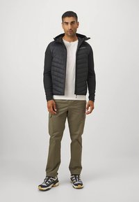 Black puffer vest with long sleeves, a hood, and a zip front. Worn over a white t-shirt and paired with olive cargo pants and hiking shoes.