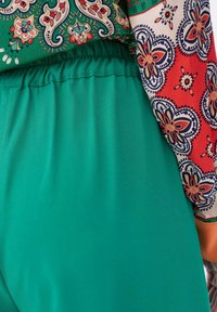 Turquoise broek met een elastische tailleband, gecombineerd met een mouw met een patroon in rood, groen en crème met bloemenontwerpen.