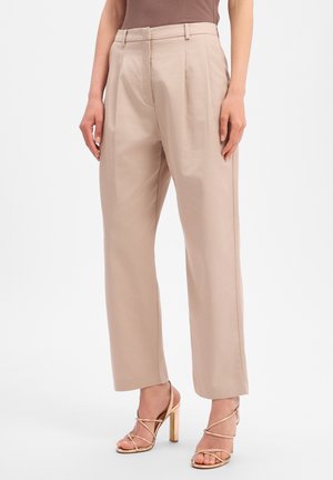 Vrouw draagt beige broek met hoge taille en wijde pijpen en gouden sandalen met hoge hak en bandjes tegen een witte achtergrond.