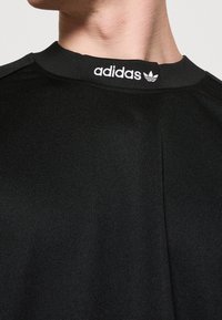 Czarna koszulka Adidas z bliska, ukazująca białe napisy "adidas" i logo tridrożka na obszarze kołnierzyka z przodu.