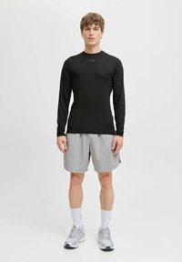 Junger Mann steht in einem schwarzen Langarmshirt, hellgrauen Shorts, weißen Socken und grauen Sportschuhen vor einem schlichten weißen Hintergrund.