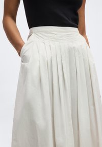 Naine kannab valget volditud seelikuga, millel on taskud, ning musta varrukateta tippu, käed taskutes.