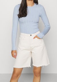 Femme portant un haut à manches longues côtelé bleu clair et un short en denim écru taille haute à ourlet brut, une main dans la poche.