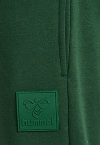 Grüner Stoff mit einem aufgenähten quadratischen Patch mit dem Hummel-Logo und Markennamen im erhabenen Design.