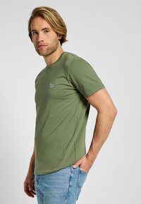 Oliwkowy t-shirt wykonany z dzianiny, z okrągłym dekoltem, krótkimi rękawami i małym czarnym logo po lewej stronie klatki piersiowej.