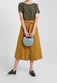 Mujer con una blusa de estampado de leopardo en verde oliva, pantalones anchos de pierna ancha color mostaza, mocasines negros con tacón, sosteniendo un bolso redondo azul.