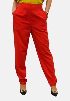 Donna che indossa pantaloni rossi a vita alta e sagomati, top giallo, tacchi neri con punta a punta e dettaglio dorato, mani nelle tasche e braccialetti al polso.