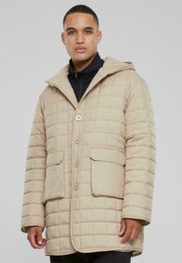Beige, quiltet, hættejakke med store frontknapper og to påsyede lommer. Glat stof med horisontale og vertikale syninger.