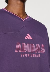 Sudadera morada con un logo rosa y el texto "ADIDAS SPORTSWEAR". El cuello tiene un acento rosa y un detalle de diseño texturizado.