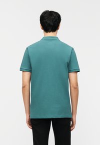 Homme debout, de dos, portant un polo à manches courtes turquoise et un pantalon noir, devant un fond blanc uni.