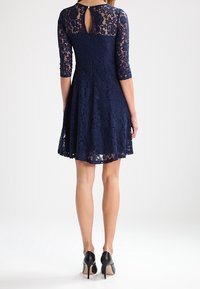 Marineblauwe kanten jurk met driekwartmouwen, uitlopende rok, een sleutelgatdetail op de rug en een gestructureerde stof. Gecombineerd met zwarte hoge hakken.