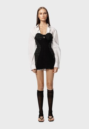 Black knitted corset dress with structured bodice, white oversized shirt, sheer black stockings, and black sandals. Round button detail visible.