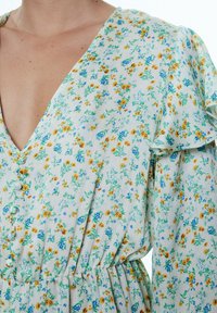 Bloemenjurk met een V-hals en korte ruffle-mouwen. Gemaakt van een lichte stof, met gele en blauwe bloemen op een lichtgroene achtergrond.