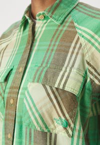 Zelená károvaná flanelová košile se zapínáním na knoflíky, náprsními kapsami a logem North Face v tyrkysové barvě na dolní kapse.