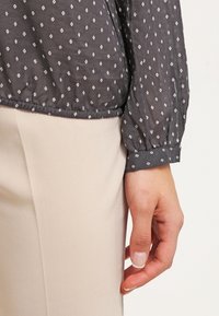 Blusa a maniche lunghe grigia scura con un sottile motivo a diamante, orlo elastico e polsini con bottone, abbinata a pantaloni beige chiaro tailored.