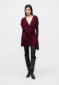 Burgundiinipunane rüütelkleit kihiliste volditud detailidega, V-kaelusega, pikkade varrukatega ja taskutega, mille juurde on kombineeritud mustad põlvikud neutraalse taustaga.