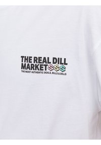 Valkoinen puuvillapaita, jossa on musta graafinen teksti "THE REAL DILL MARKET" värikkäillä dollarimerkeillä ja tunnuksella alapuolella.